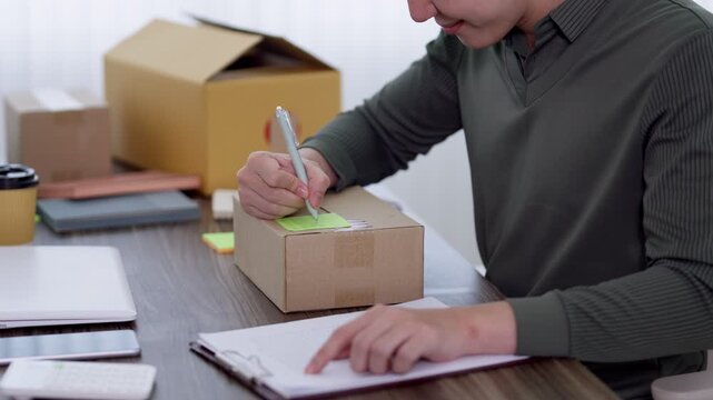Asian male entrepreneur writing address label on parcel while checking shipping order document at home office desk
