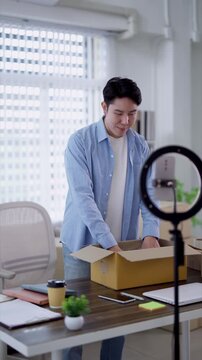 Young Asian entrepreneur unpacking product from parcel box while filming livestream for online store in home office