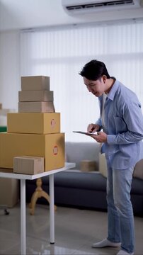 Young Asian entrepreneur checking digital tablet for shipping orders beside stacked parcels in modern home office