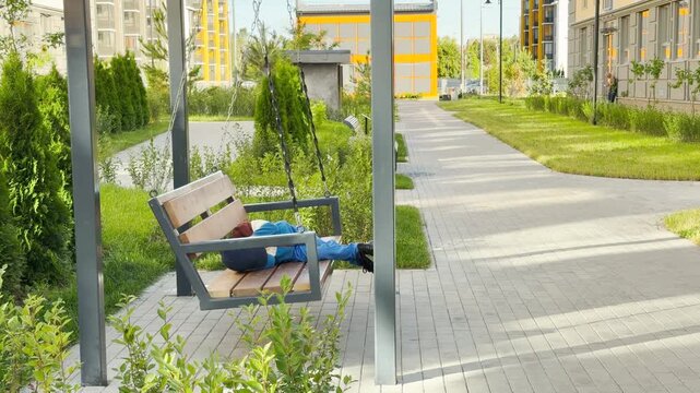 A young boy relaxes and rests comfortably on a hanging wooden swing in a sunny park. This peaceful scene evokes a feeling of calm, cozy contentment and sweet leisure.