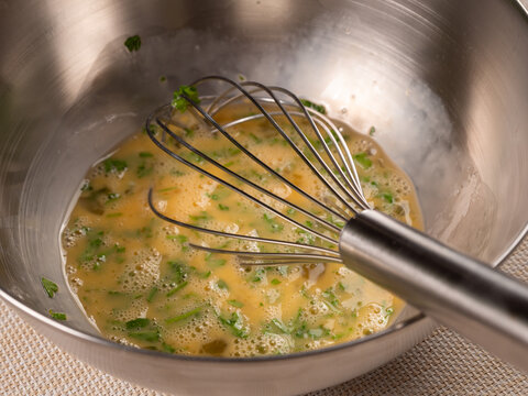 A whisk blends golden beaten eggs with flecks of fresh parsley in a shiny stainless steel bowl. The close angle shows the frothy texture forming during meal preparation.