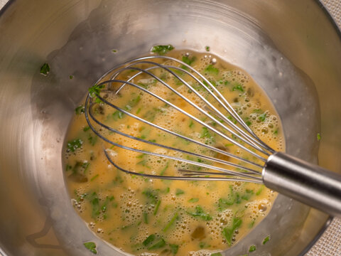 A metal whisk stirs beaten eggs mixed with chopped fresh parsley inside a stainless steel mixing bowl. Small bubbles form on the surface as the egg mixture is prepared for cooking.