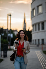 Fototapeta premium Woman smiling, walking with smartphone and coffee urban sunset