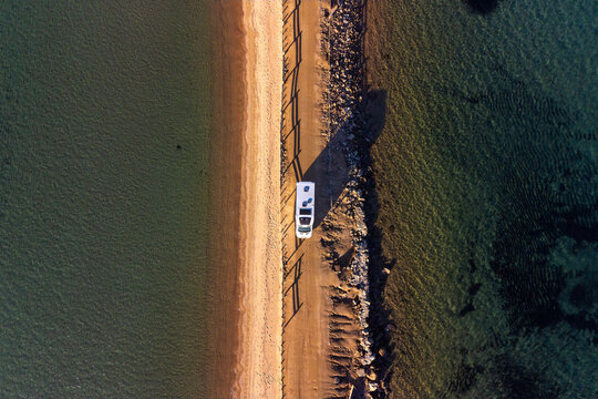 Aerial view of a camper van parked on the narrow sandy isthmus leading to Seagull Island, surrounded by clear turquoise waters in Porto Pollo, Sardinia, Italy.