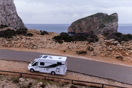 Aerial view of a white camper van parked on a coastal road near the rocky cliffs of Capo Caccia with the Mediterranean Sea in Alghero, Sardinia, Italy.
