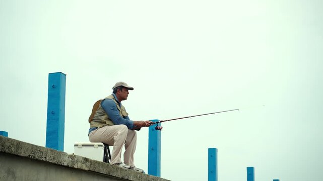 Happy senior man sitting by the sea and fishing with a rod after retirement. Active aging, healthy longevity, outdoor leisure, fulfilling retirement. Life insurance and financial security concept.
