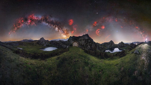 View of the Lakes of Covadonga under a vibrant Milky Way galaxy and starry night sky, featuring a small stone building amidst grassy mountain terrain in Asturias, Asturias, Spain.