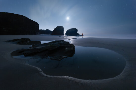 View of As Catedrais beach at night with a lone person standing on the sand near tide pools under a bright moonlit sky in Galicia, Spain.