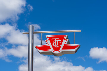 Naklejka premium Toronto, ON, Canada - August 17, 2026: A red and white exterior sign featuring the Toronto Transit Commission (TTC) logo against a blue sky with white clouds, which marks a public transit subway or ra