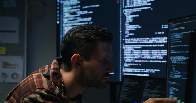 Caucasian male software engineer types code at a dim office desk, debugging a web app while monitoring logs and security alerts on multiple screens.
