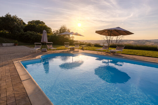View of a swimming pool at sunset with lounge chairs and umbrellas overlooking the rolling hills of the Marche region in Macerata, Marche, Italy.