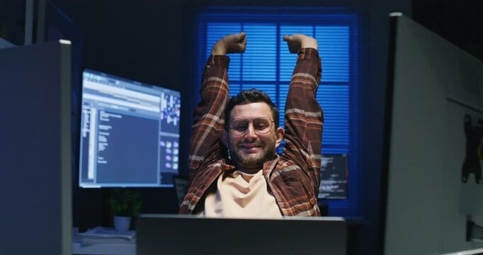 Caucasian male programmer sits slumped at his desk after a late deployment, monitors dimmed, success mixed with stress and fatigue.