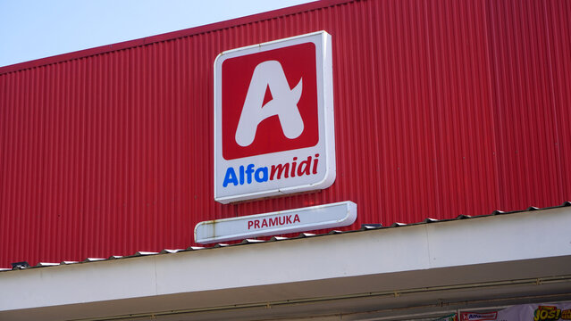 A red facade of an Alfamidi convenience store branch on Pramuka Street in Kudus, Central Java, Indonesia, featuring its official logo, signboards, and promotional posters on a bright sunny day.