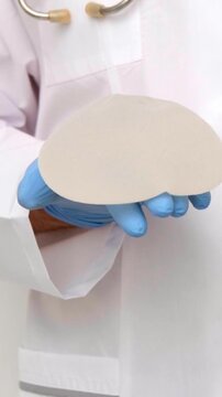 A medical professional in blue gloves presents a silicone breast implant in a clinic room, plastic surgery and aesthetic medicine healthcare services, patient consultation and examination.
