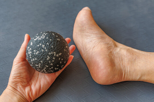 Close up of hand holding fascia massage ball near bare foot on exercise mat, foot massage and plantar fasciitis relief concept