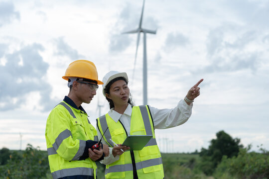 Female wind energy engineer pointing toward turbines while discussing field operations with a technician, representing renewable power teamwork, inspection, planning and maintenance.