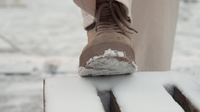 Low-angle close-up of brown boot pressing into snow-covered slatted bench, snow compressing under sole, tread detail visible, with cold winter light and textured outdoor surface, with snow, boot