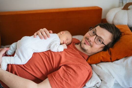 Father and baby son bonding closely at home, with the newborn sleeping soundly on dad's chest while the father looks at the camera, capturing a tender moment