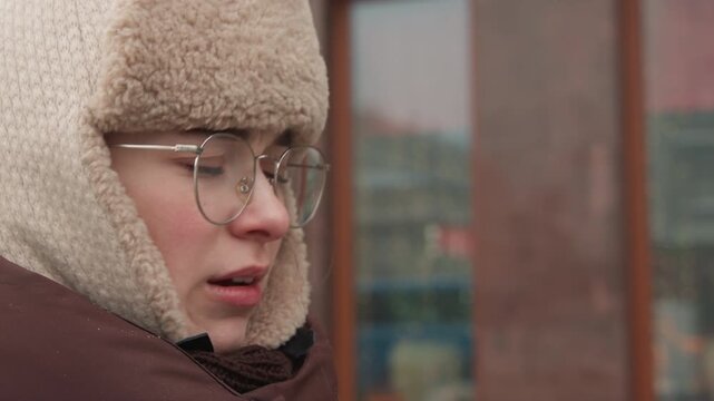 Young person wearing thick fleece hat with ear flaps, glasses, heavy coat, and scarf cups hands near mouth to warm breath; faint vapor, reddened cheeks, snowy street, soft daylight, quiet winter mood