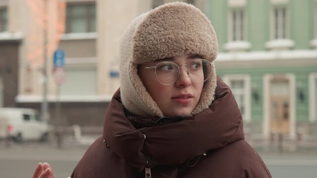 Rear quarter portrait of woman wearing knit trapper hat and padded winter coat, turning head and looking right while snow falls and blurred vehicle moves through urban traffic in cold overcast light