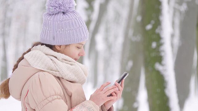 Beautiful girl in jacket and hat with a bright scarf reads the news on the Internet on a smartphone on a background of a winter city park.