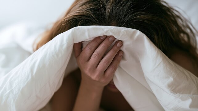 Person hiding face with hands under blanket in a bedroom during morning hours