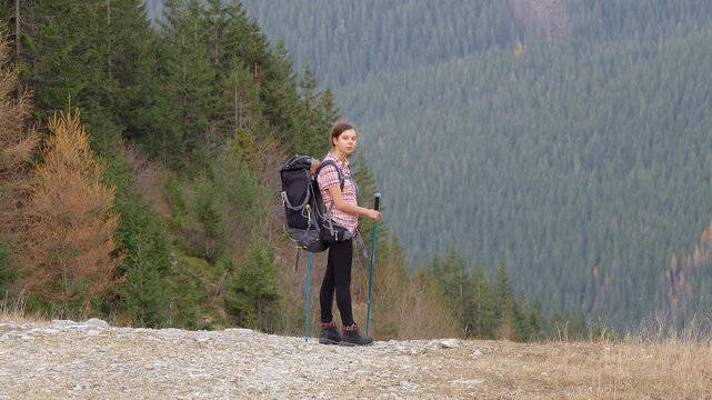 Young girl with scandinavian sticks and backpack on the background of the mountains. Green coniferous forests on the mountainside.