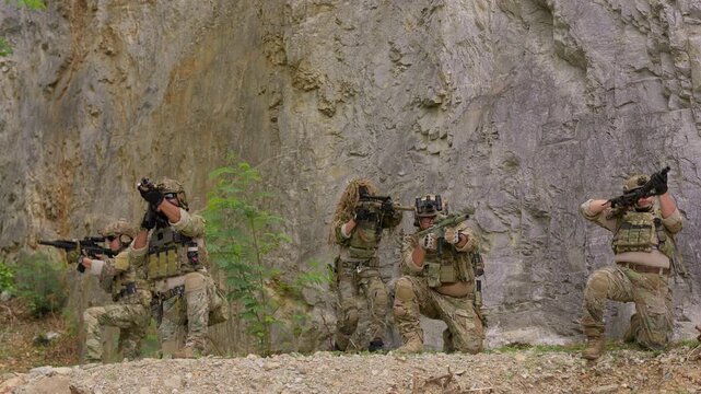 A special forces team conducts a military operation, with soldiers aiming rifles from defensive positions against a rugged rock face background.