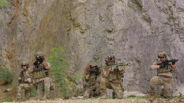 A special forces team conducts a military operation, with soldiers aiming rifles from defensive positions against a rugged rock face background.