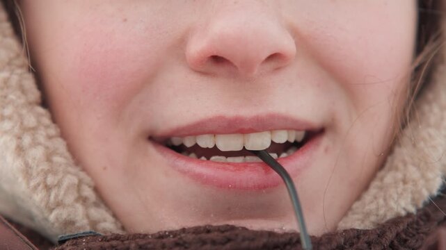 Tight frontal close-up of lower face with fleece hood and scarf, mouth open as thin black cord rests between lips and teeth; visible action shows careful adjustment in cold outdoor light