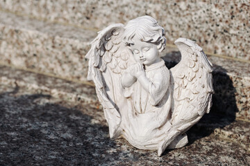 Fototapeta premium White praying angel statue rests outdoors. Decorative wings surround peaceful child figure. Religious ornament stands on stone surface. Memorial sculpture forms spiritual background.