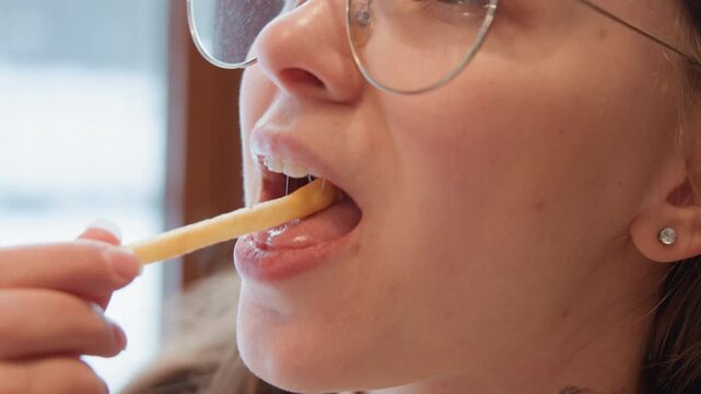 Close profile view of young woman in round glasses lifting french fry to lips by bright window, gentle smile, soft jaw movement, casual indoor snack scene in natural daylight, with woman, glass, fry