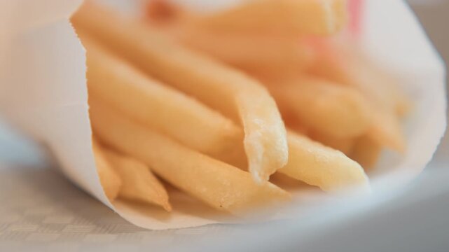 Tight low-angle close-up showing golden French fries filling white paper sleeve, with brief hand motion and settling shapes, warm diffused light, crisp texture, and casual snack appeal