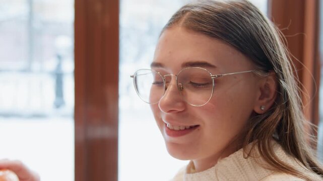 Young woman with clear glasses lifts sandwich toward mouth while smiling beside snowy window, calm indoor mood with hand movement, soft light, relaxed posture, and clear hunger anticipation