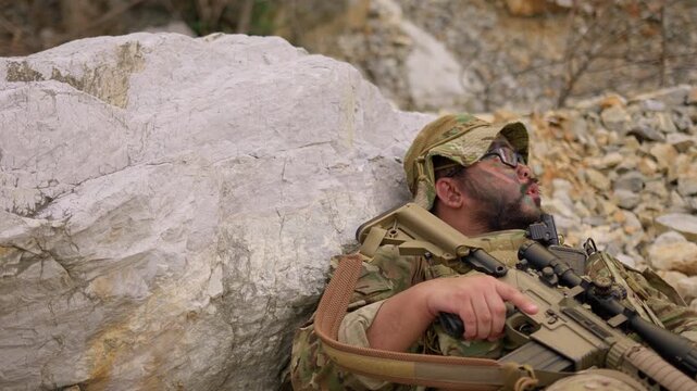 An exhausted soldier with face paint rests behind a rock, holding his rifle and catching his breath during a tactical military mission in a rugged environment.