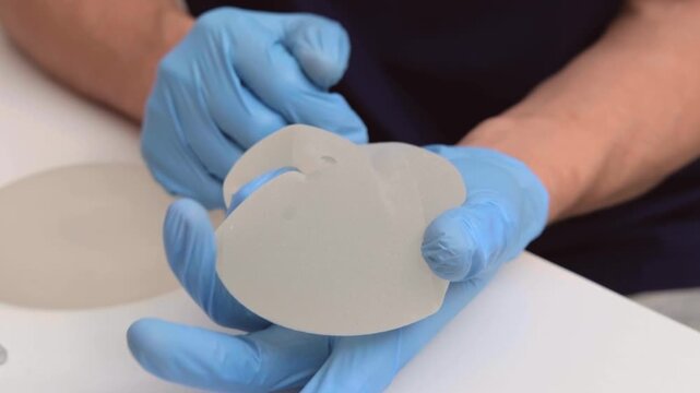 Surgeon in blue gloves pressing and testing a silicone breast implant on a table, healthcare advertising, plastic surgery, medical examination.