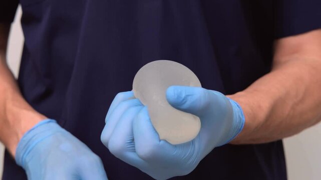 Surgeon holding a silicone breast implant and demonstrating its softness and shape, healthcare advertising, plastic surgery, medical equipment.