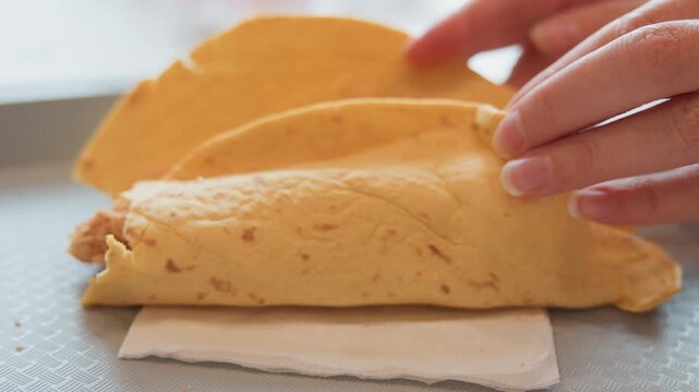 Close view of hands folding thin golden crepe over shredded protein and small vegetable pieces on paper lined countertop, showing careful lifting, tucking, and shaping motion during food preparation