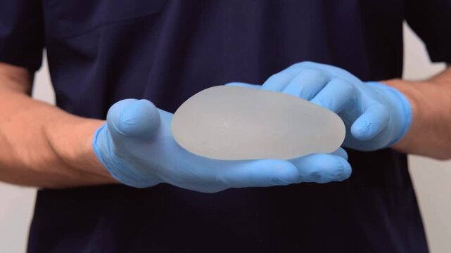 Surgeon hands in blue gloves demonstrating a flexible silicone breast implant, healthcare advertising, plastic surgery, medical equipment.