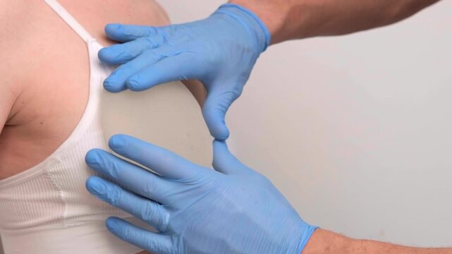 Surgeon in medical gloves checking breast implants on a female patient in a clinic, healthcare advertising, plastic surgery, physical examination.