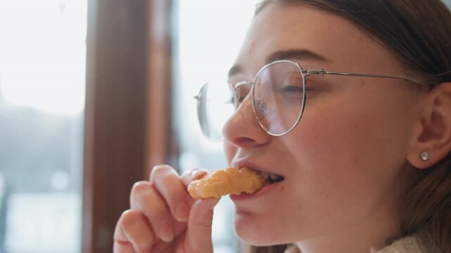 Young woman savoring fried snack with satisfied expression in sunlit cafe while seated by tall window, hand lifting bite near mouth, soft daylight shaping relaxed face and calm indoor mood