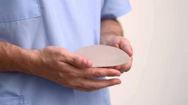Plastic surgeon in scrubs demonstrating silicone breast implant softness and elasticity, aesthetic medicine clinic, augmentation mammoplasty patient consultation.