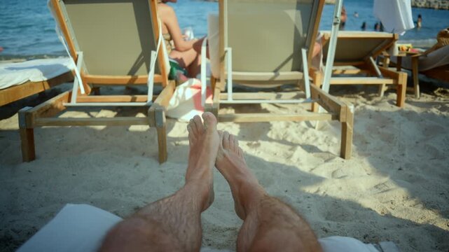 Person enjoying summer vacation, lounging on a beach chair with sandy feet, looking towards the ocean and other beachgoers