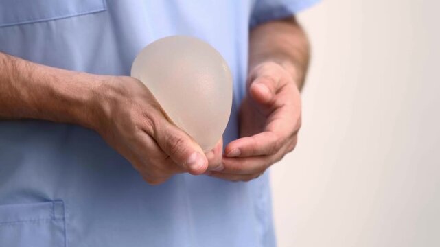 Surgeon squeezing and twisting a breast implant to show durability in scrubs, plastic surgery clinic, aesthetic medicine and augmentation mammoplasty presentation.