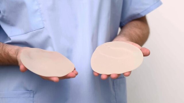 Plastic surgeon in surgical scrubs holding two breast implants for augmentation mammoplasty comparison, aesthetic medicine clinic, patient consultation presentation.