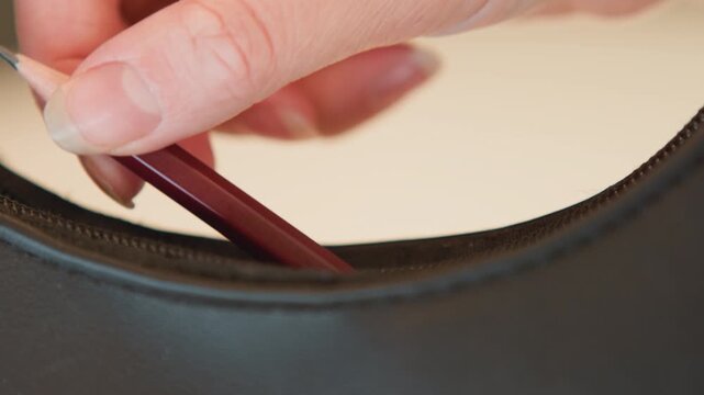 Close view shows fingertips pressing thin tool into narrow gap along curved dark leather upholstery edge, creating slight indentation, tension, and careful probing beside stitched seam