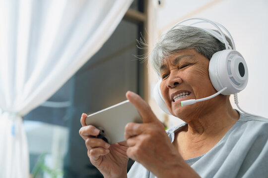 Elderly Asian woman relaxing on a sofa at home, wearing headphones while enjoying games on her smartphone, modern living among older adults.