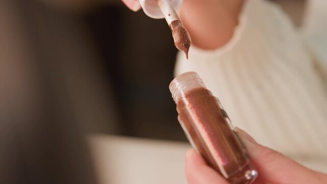 Close-up view of hands holding false nail tip while brush lifts above surface with clear polish dripping from bristles back toward bottle, showing wet shine, careful grooming, and salon style beauty