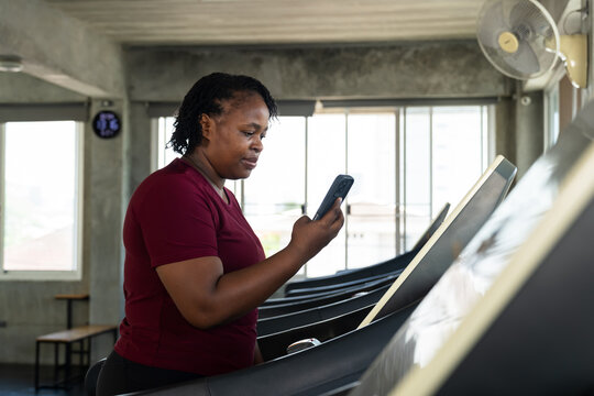 Plus size African American woman holding smartphone near treadmill screen in modern gym, active fitness lifestyle photo for cardio routine, wellness progress, body confidence motivation and self care.