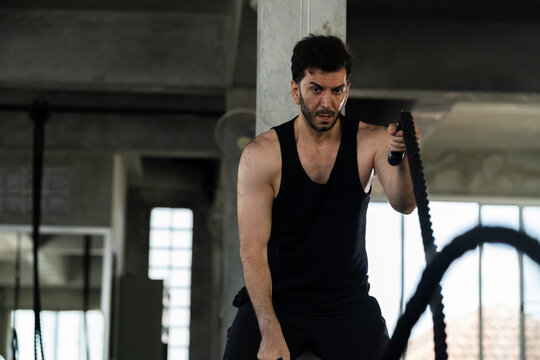 Adult Caucasian man using battle rope in modern gym, male athlete leaning forward during functional cardio strength training, high intensity endurance workout for gym membership, men fitness ads